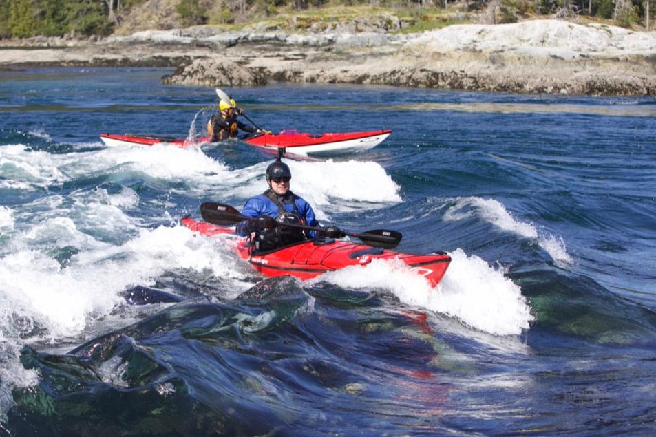 Two people kayaking in rough waters, with one kayak splashing more water than the other, indicating a dynamic and exciting kayaking experience.